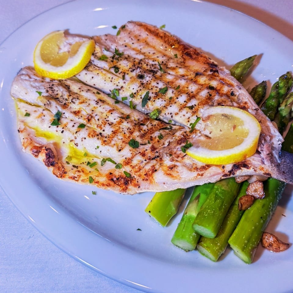 Grilled fish fillets with lemon slices, fresh herbs, and asparagus on a white plate.